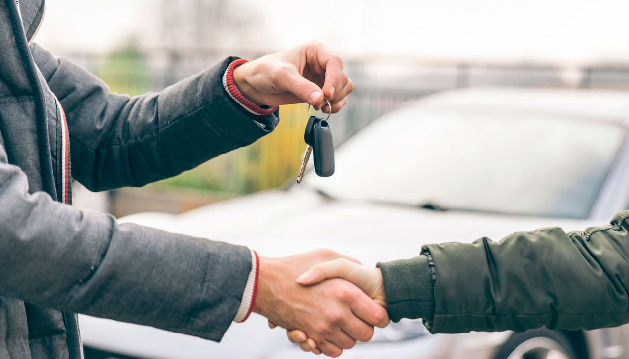 Two people shaking hands in front of a car as one of them hands over the car keys.