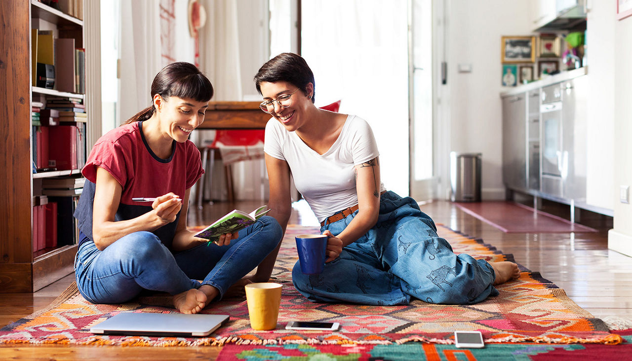 Two young women sitting on a living room floor, creating a list with pen and paper