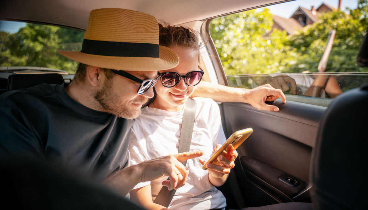 Young cheerful happy couple of travelers sitting in car looking at smartphone screen, travel comfort safety taxi