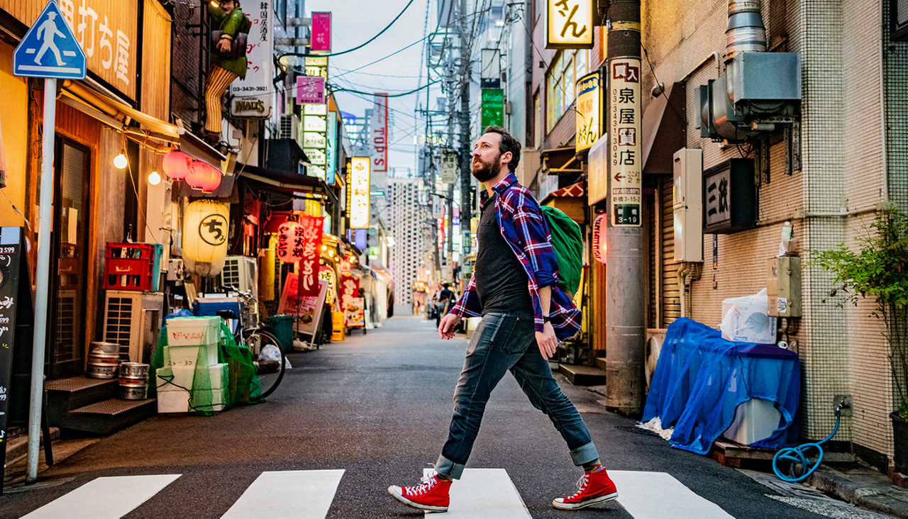 A man crosses a narrow street packed with bright signage. 