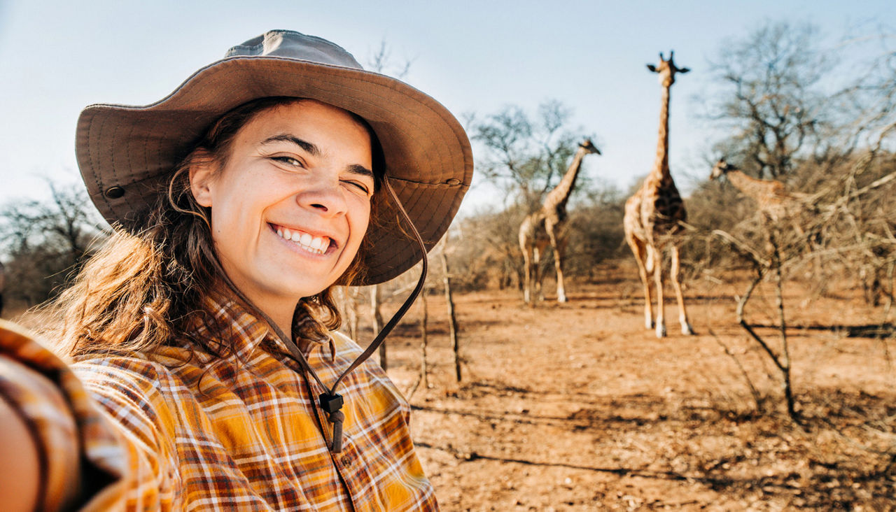 Portrait of a happy female tourist enjoying the nature reserve and taking a selfie with giraffes in the background