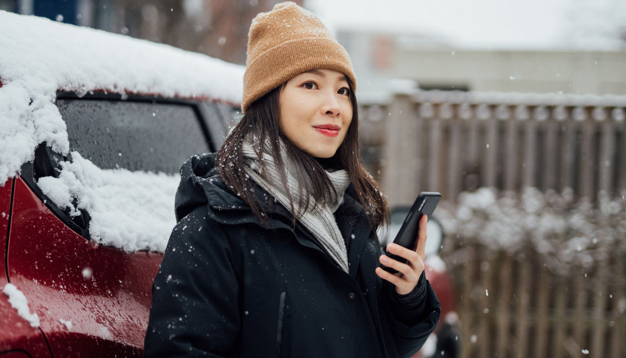 Smiling young woman in warm clothes ordering taxi with smartphone while standing in snowy weather