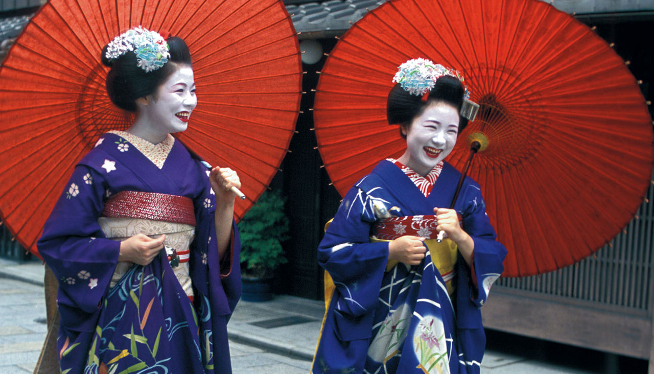 Two Geisha women dressed in blue robes with red umbrellas