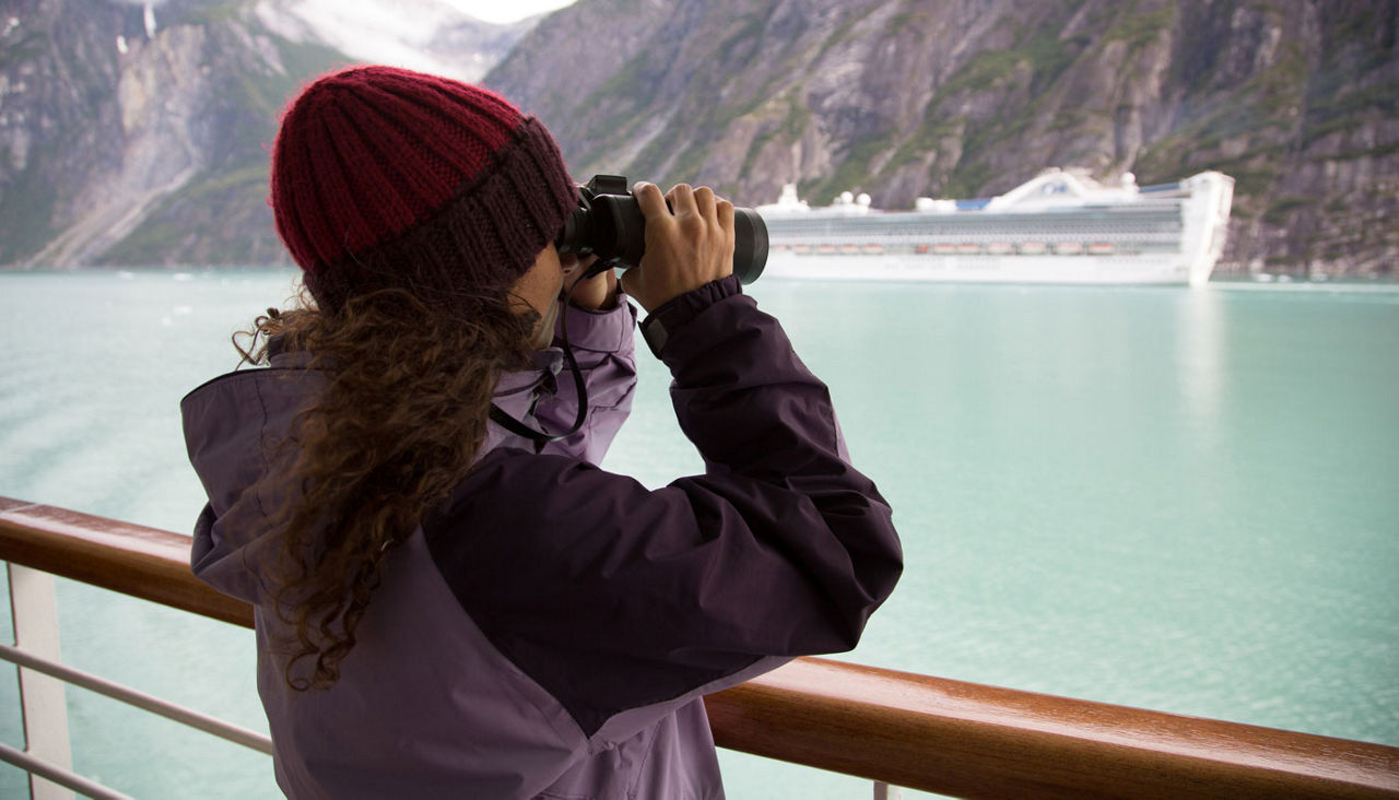 woman peering through binoculars at Alaskan scenery from cruise ship