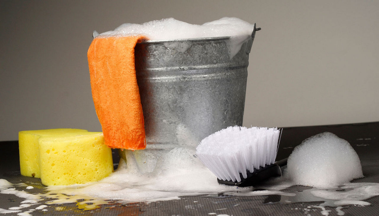 A close-up view of a bucket with soapy suds coming out the top and it is surrounded by brushes, sponges and rags.