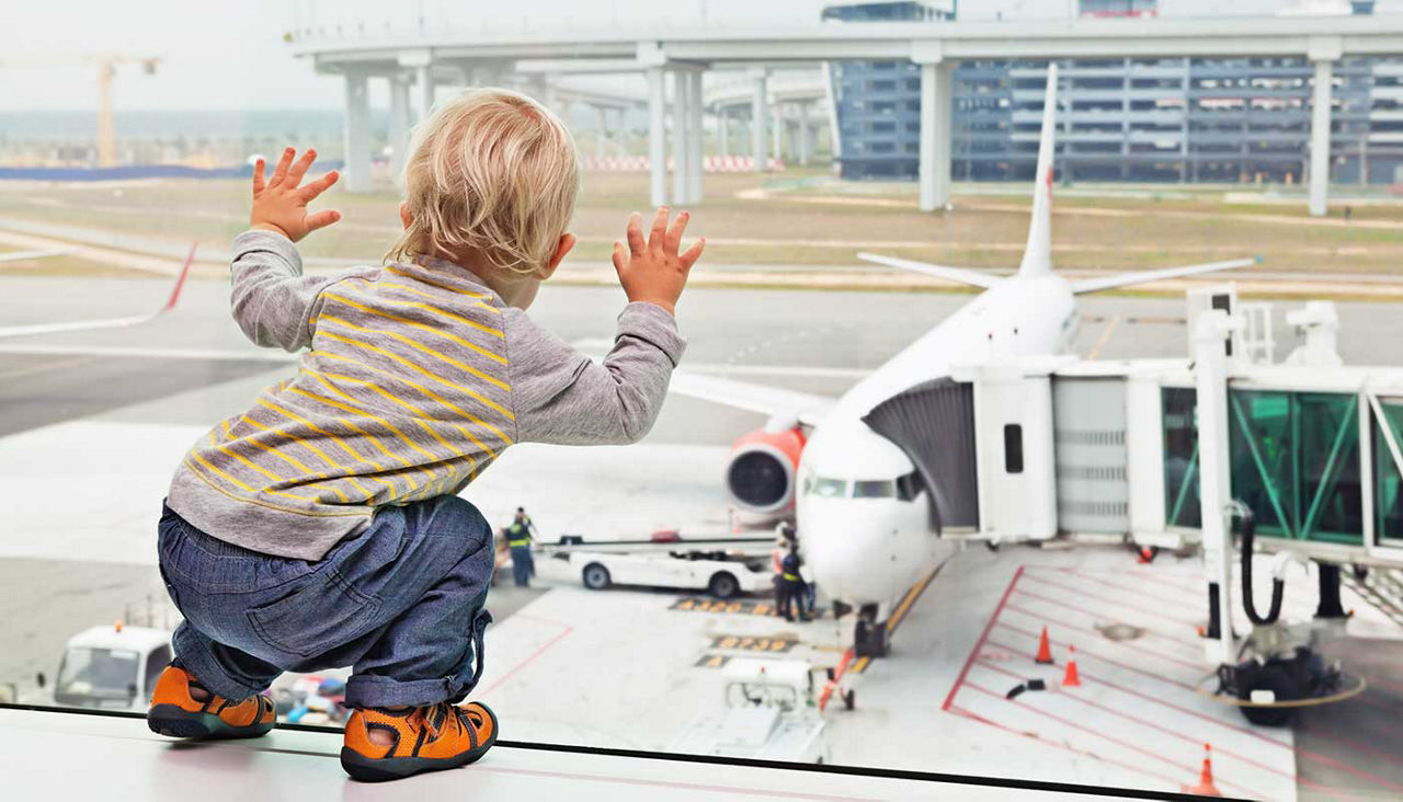 Little baby boy waiting boarding to flight in airport transit hall and looking through the window at airplane near departure gate. 