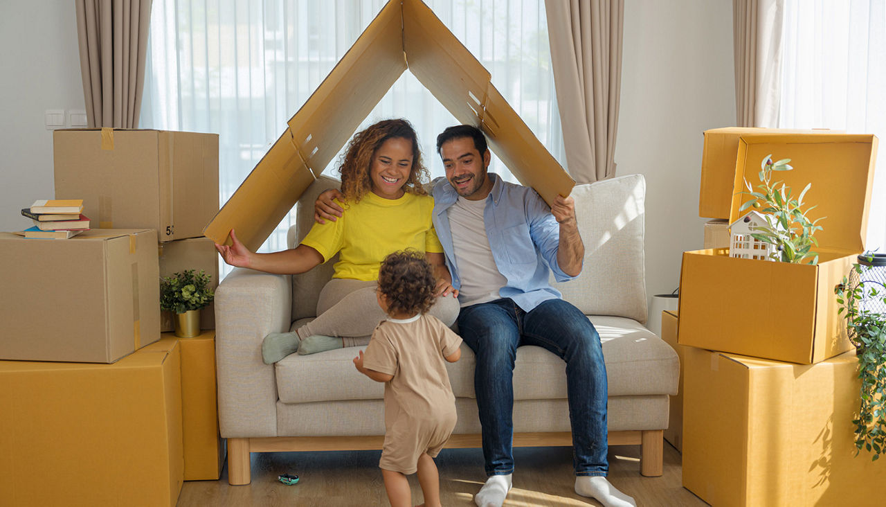 Two parents surrounded by cardboard boxes in a living room holding a broken down cardboard box over their heads and smiling at their toddler