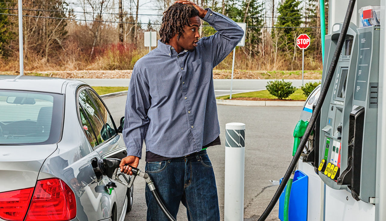 A young man putting gas into his car looking shocked at the price of the gas at the pump.