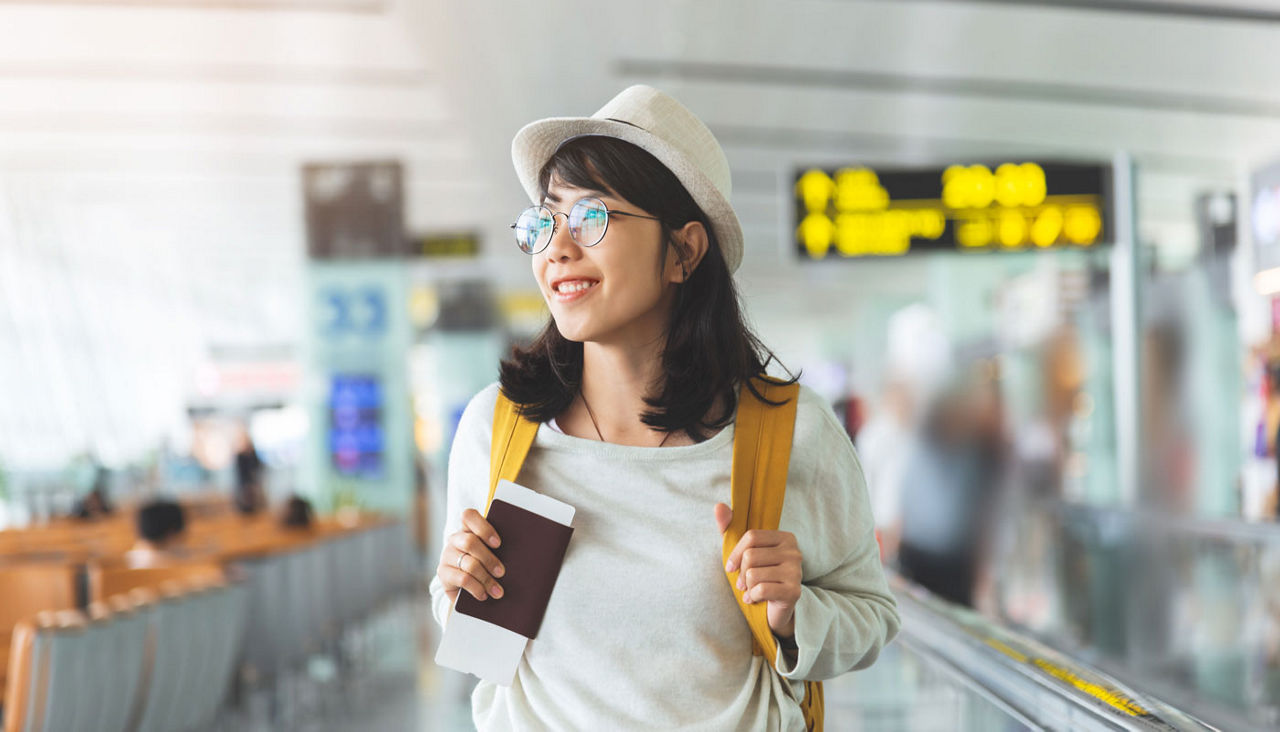 Asian woman with glasses and hat is holding airline ticket, passport at airport.