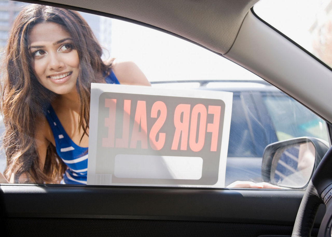 A woman looking into a car from the window with a For Sale sign visible on the window