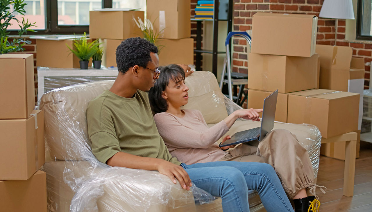 Couple moving into new home sitting on sofa and looking at laptop while surrounded by boxes 