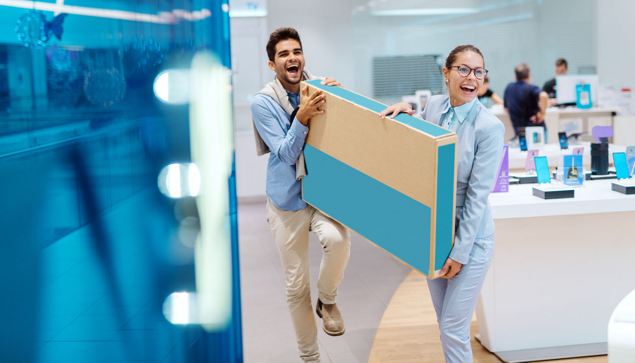 Happy multicultural couple holding box with new television in it. Tech store interior.