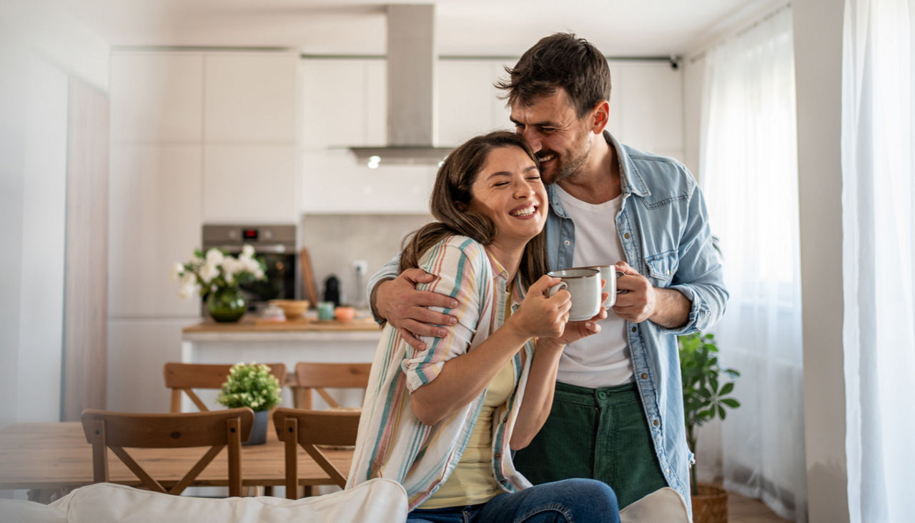 Man and woman hugging in kitchen holding coffee mugs