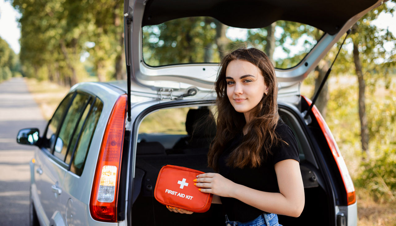 Young girl standing near open back door of silver hatchback car and shows first aid kit that must be in every car for emergency