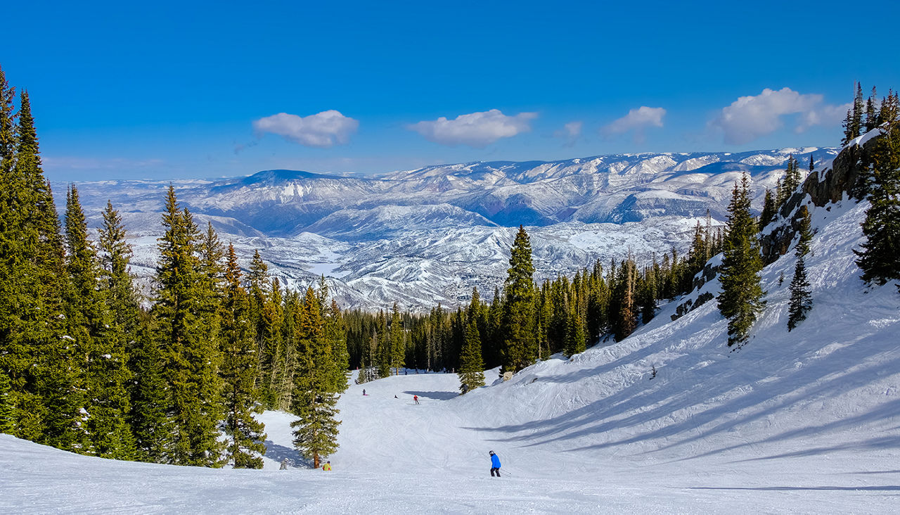 View of ski slope in Colorado ski resort on clear winter day; spruces, pine trees, blue sky and mountains in background