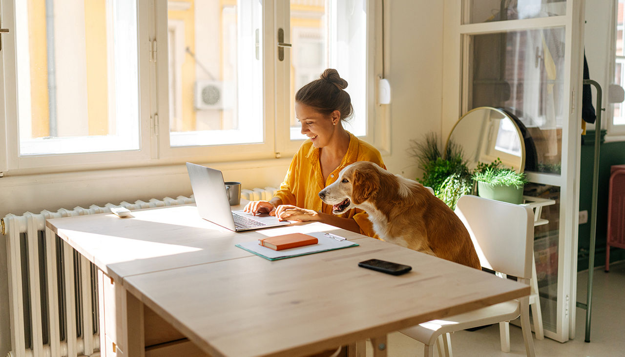 A woman smiles as she works at her laptop, a dog seated next to her at the table.