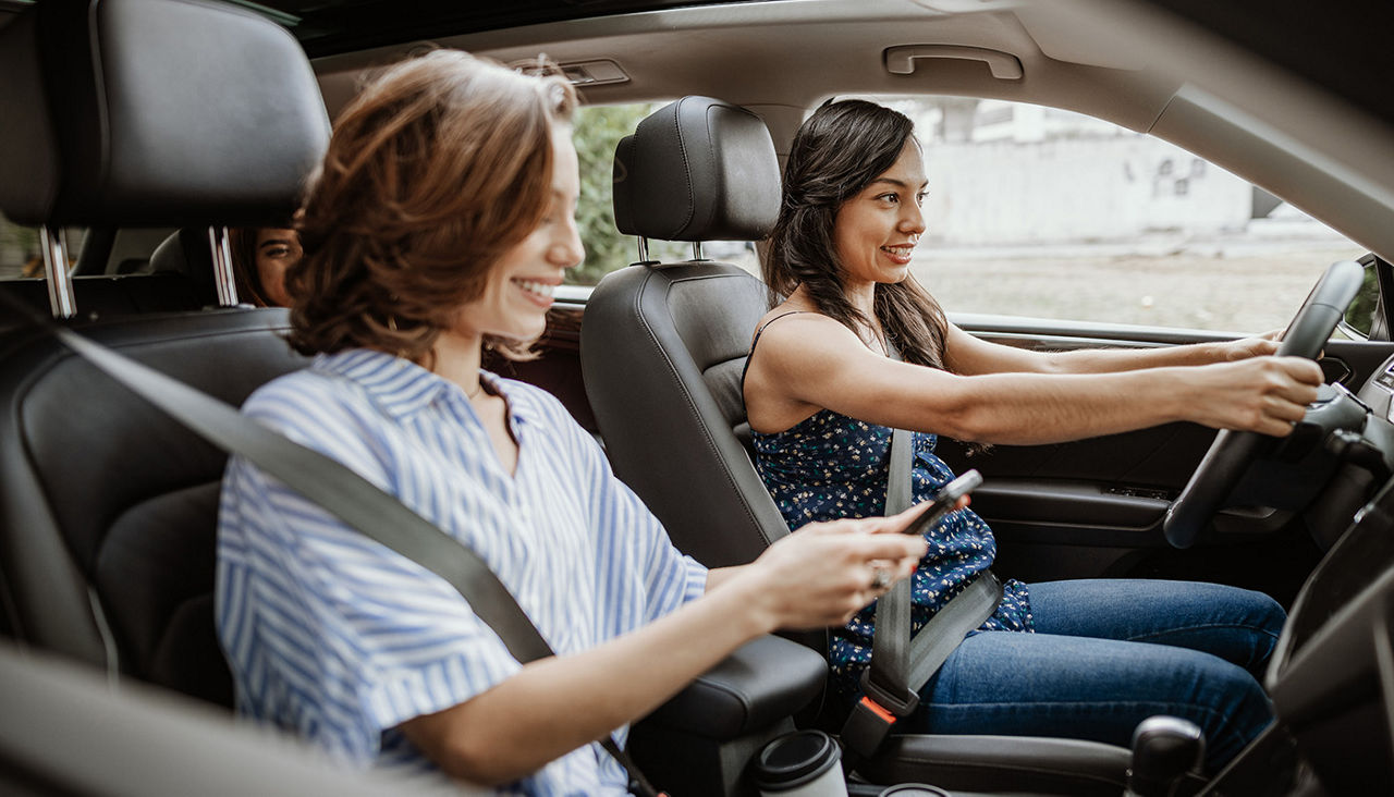 A woman in the passenger seat checks a cell phone while the woman in the driver’s seat maintains eye contact on the road ahead.