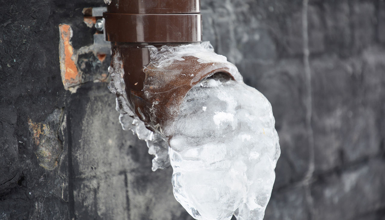 A plastic downspout, downpipe of a roof gutter system with ice and frozen water in the pipe near a house foundation in winter.