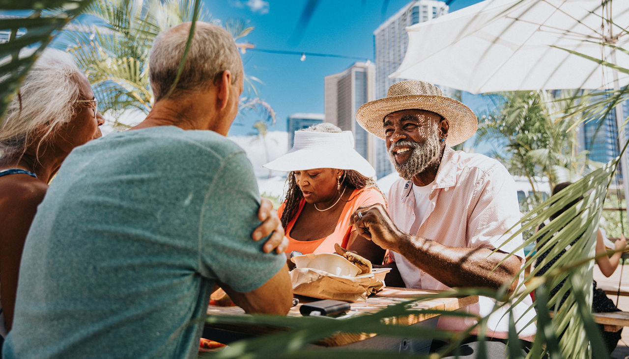 A vibrant African American couple enjoy eating local street food purchased at a farmer's market food fair while dining outdoors with friends they are visiting in Hawaii.