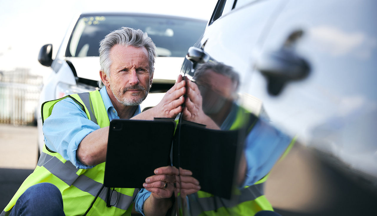 Mature male insurance loss adjuster wearing hi-vis safety vest standing in compound for damaged cars compiling insurance claim report on digital tablet