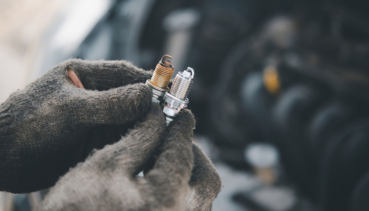 Gloved hands hold up a new and a used spark plug side by side.