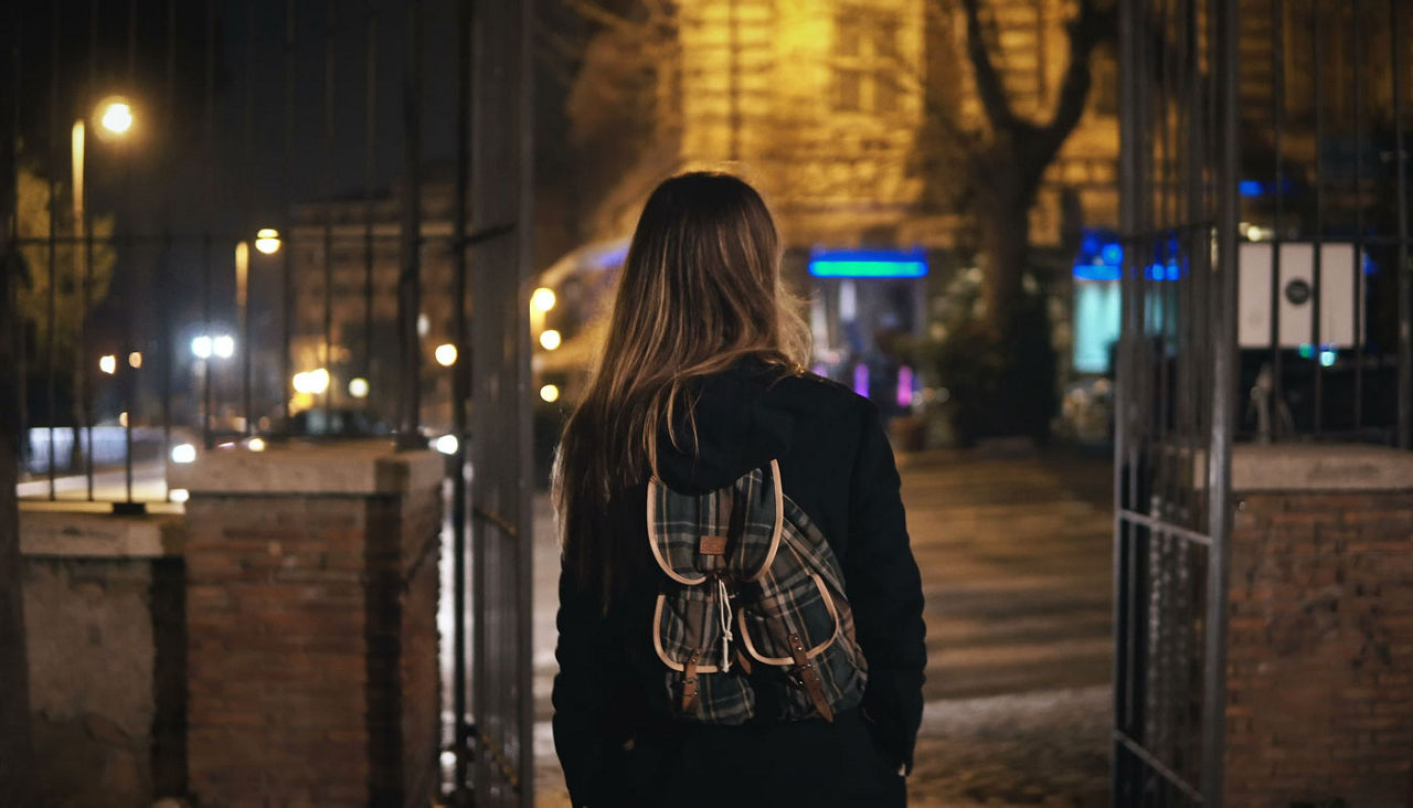 Back view of tourist woman with backpack walking through the dark park near the road late at night alone.