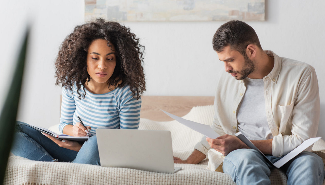 Wife and husband sitting on bed working on finances with laptop, notebook and other paperwork