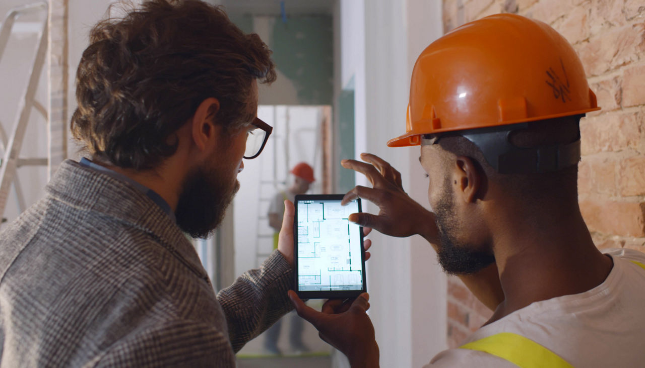 Back view of apartment owner and foreman in orange hardhat discussing blueprint on digital tablet