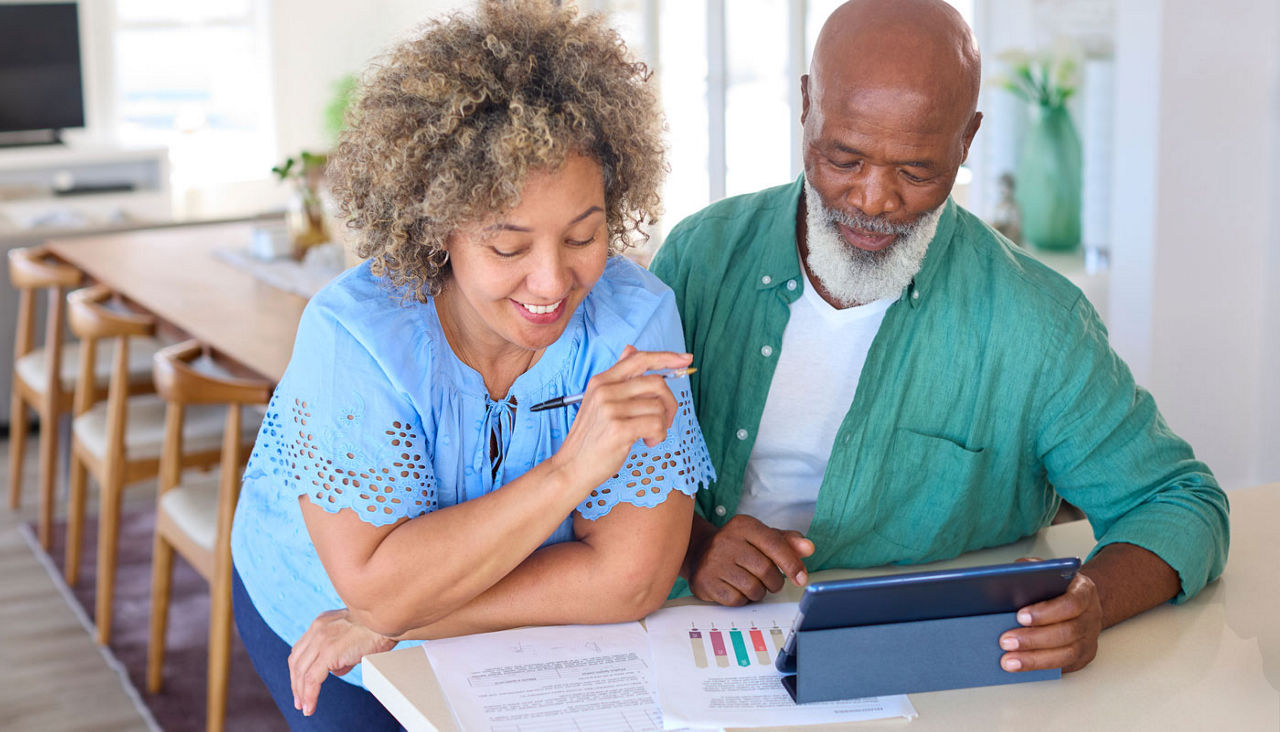 Smiling mature couple standing at kitchen counter looking at finances on tablet 