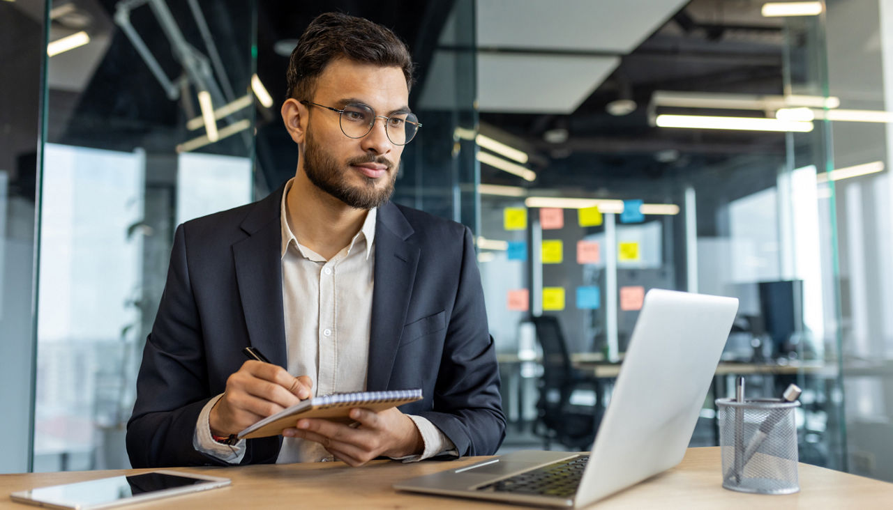 Young professional man looking at laptop and writing on notepad