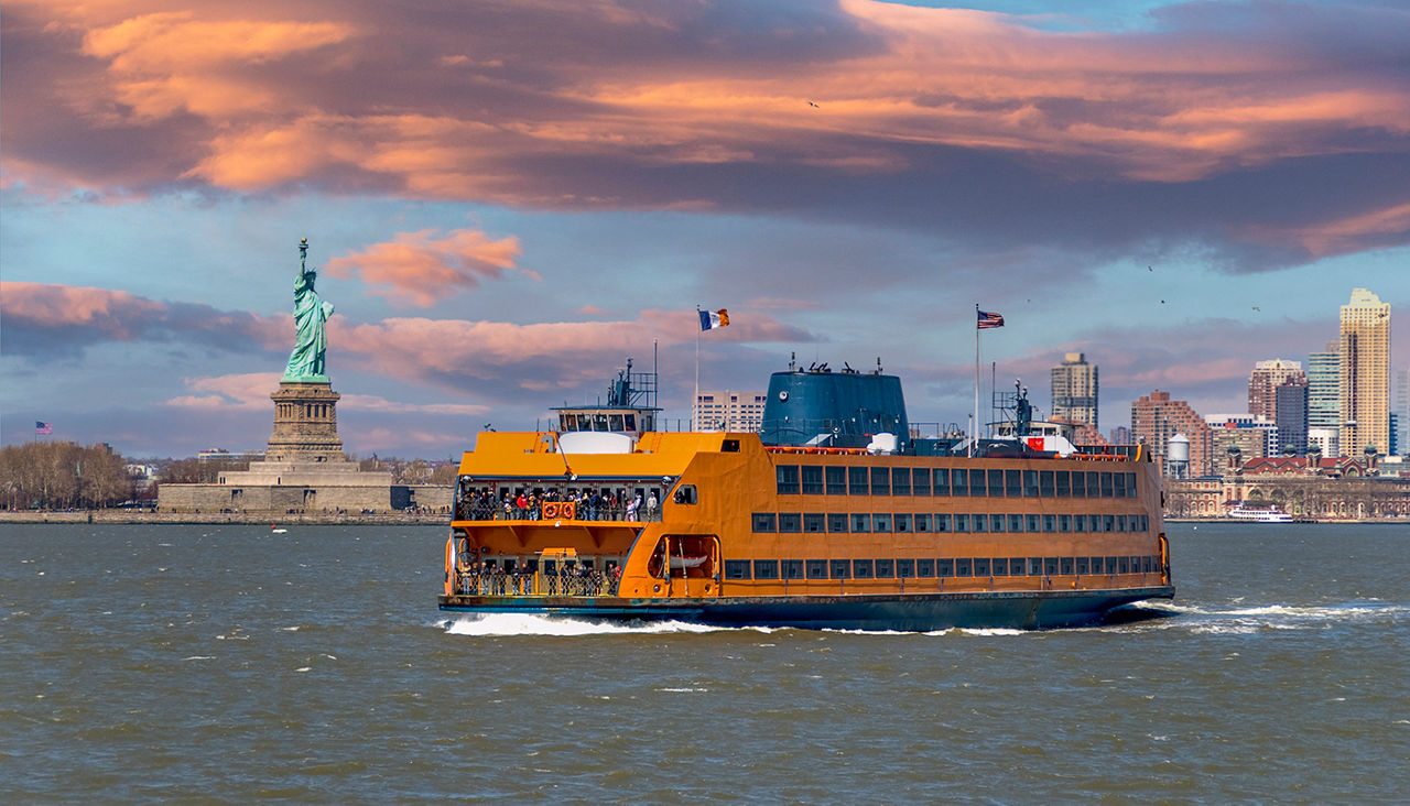 Staten Island ferry coming from Manhattan and passing by the Statue of Liberty. The Statue of Liberty and the skyscrapers of lower manhattan can be seen in the background.