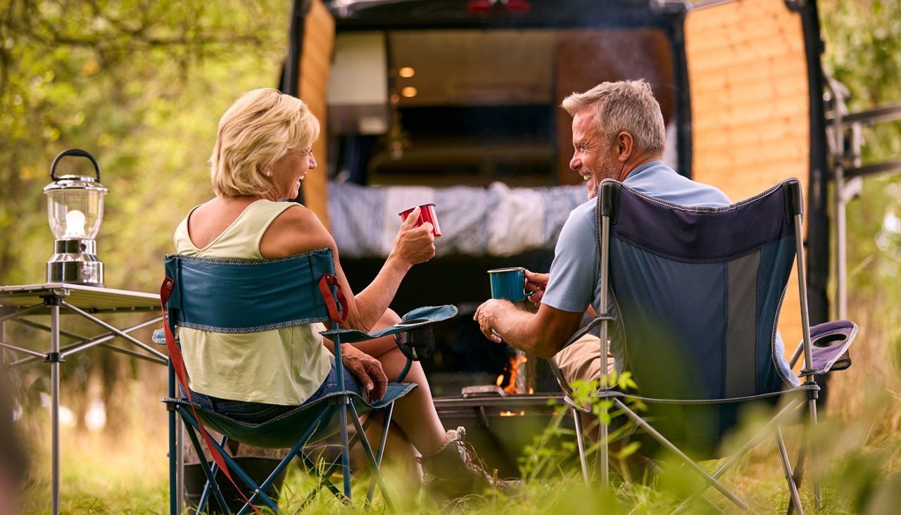 Rear View Of Senior Couple Camping In Countryside With RV Drinking Coffee By Outdoor Fire