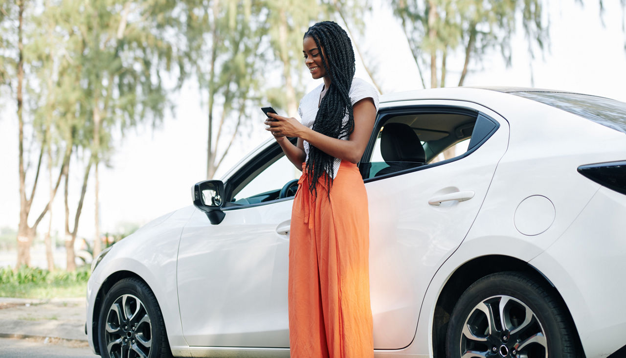 Cheerful young Black woman with long hair standing next to her car and looking at her phone