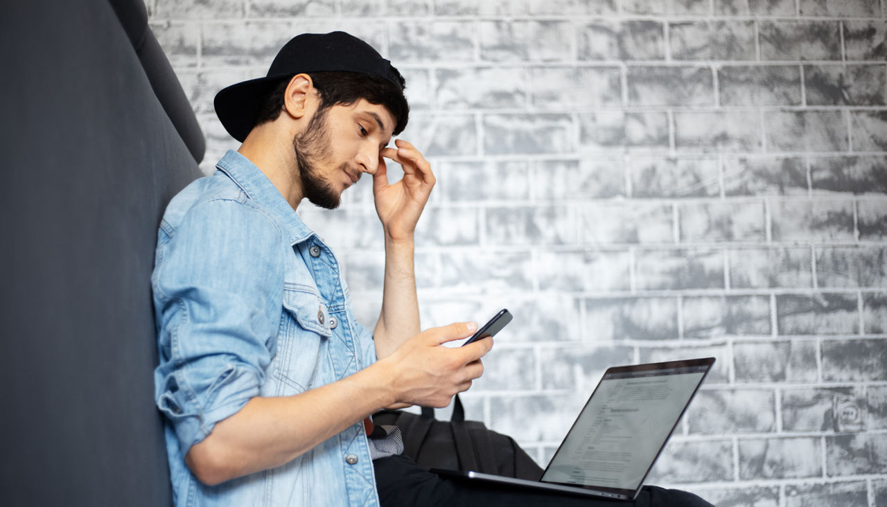Young man with concerned look on his face looking at phone while working on laptop