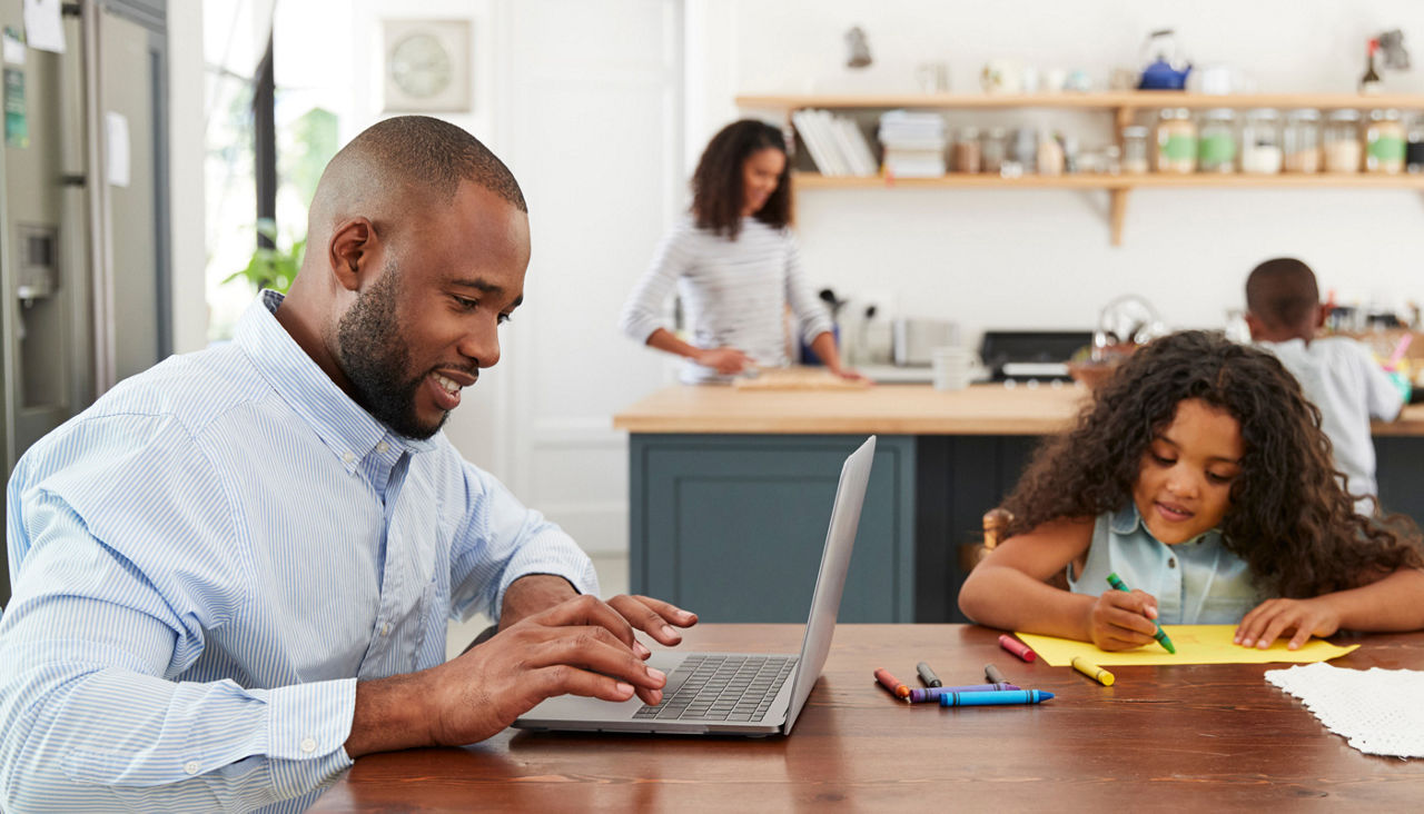 Father typing on laptop at kitchen table, his daughter coloring beside him, and his wife and young son working in the kitchen