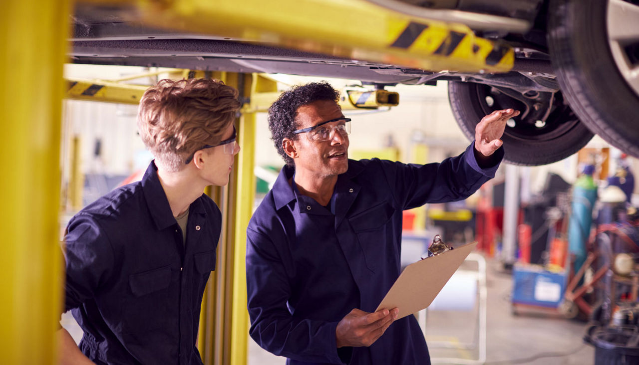 Male Tutor With Student Looking Underneath Car On Hydraulic Ramp On Auto Mechanic Course