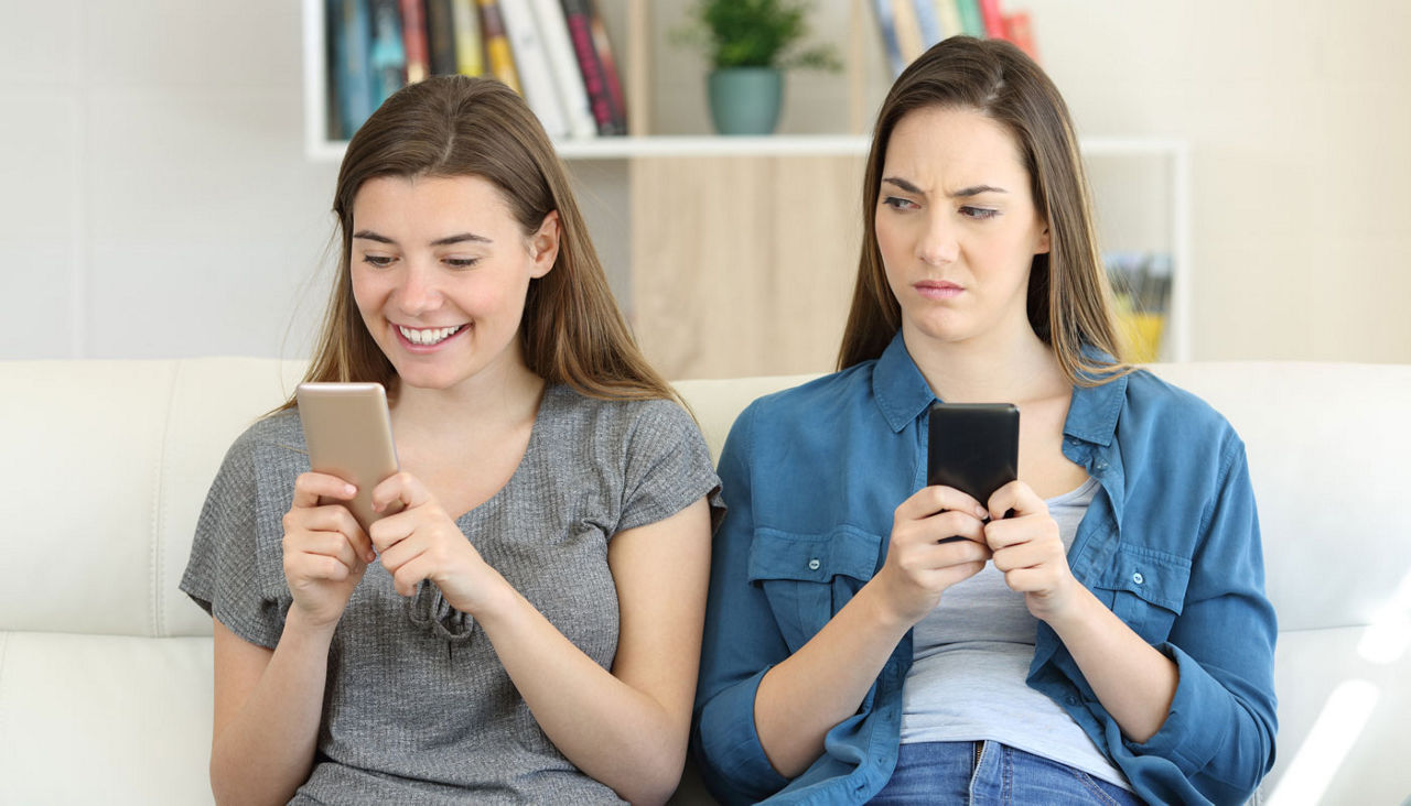 Envious woman holding phone and looking at her happy friend looking at her own phone