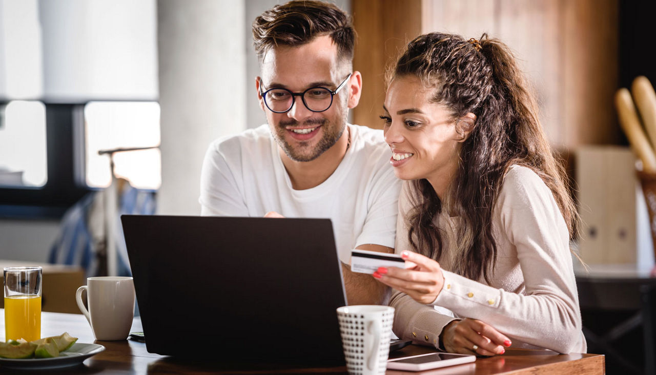 Smiling couple in kitchen using laptop and paying with credit card.