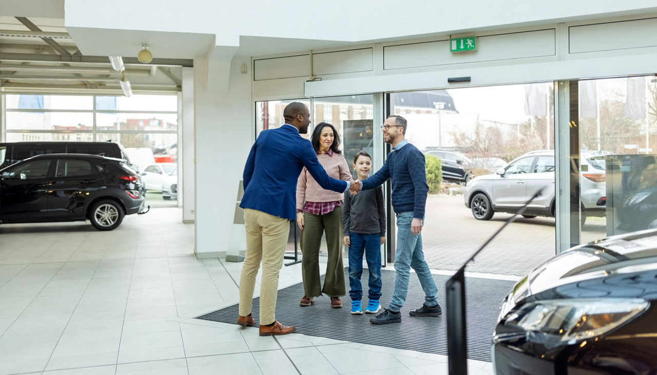 A father, mother and child walk into a car dealership and the father shakes hands with a salesperson.