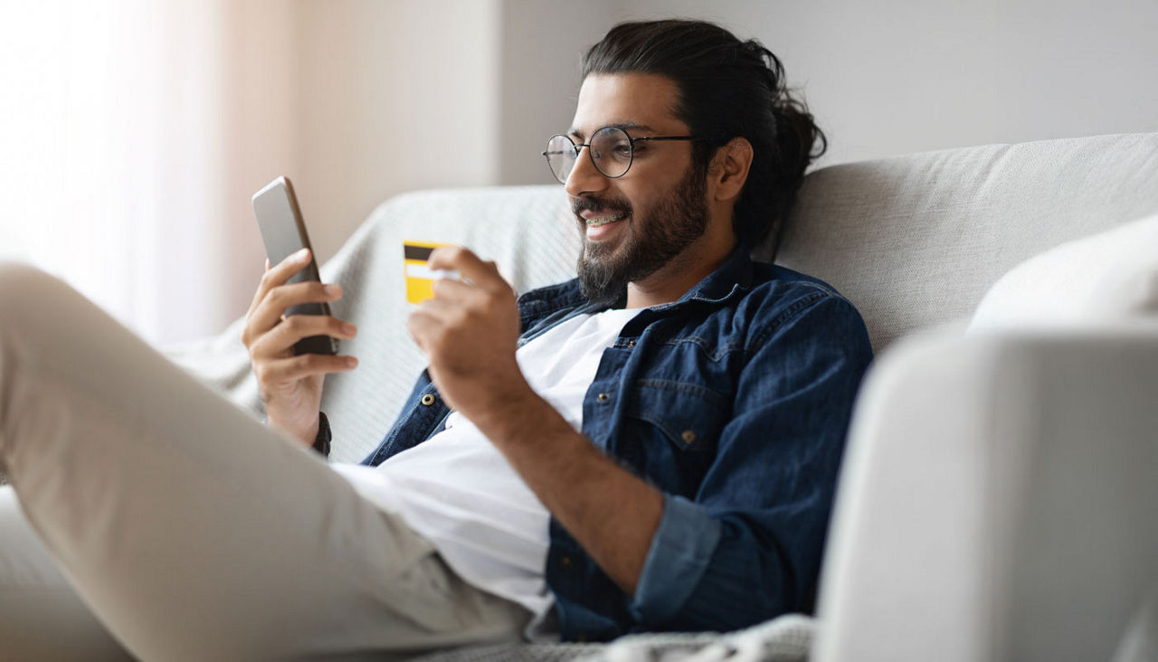 Cheerful man on sofa shopping on phone with credit card