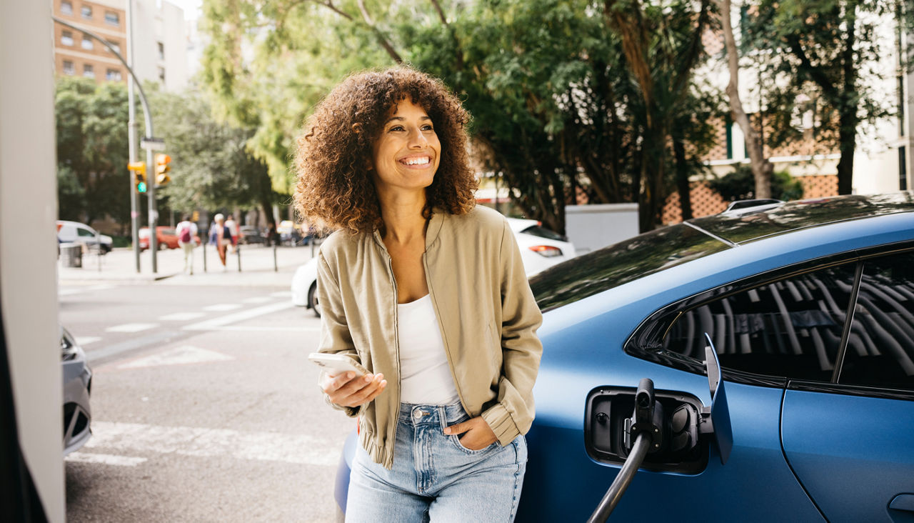 A woman is smiling as she stands next to her electric car at a charging station in a city 