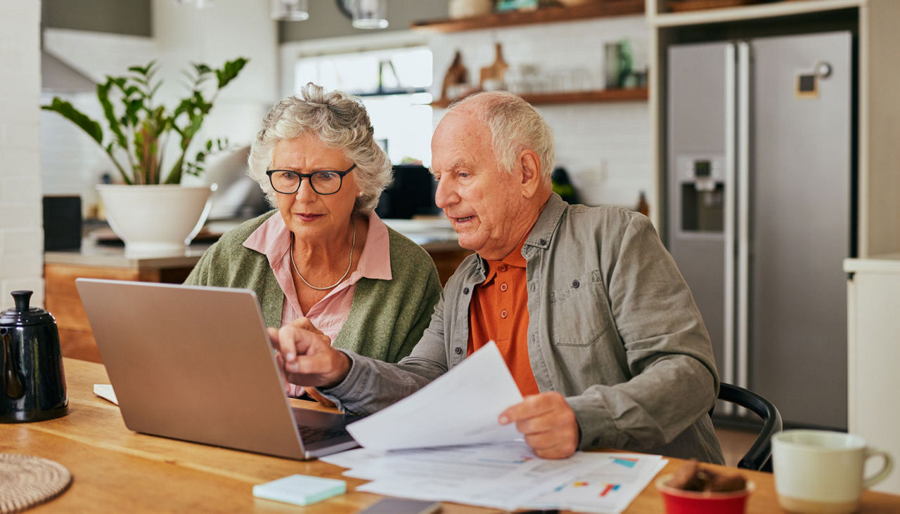Focused senior couple sitting at kitchen table, managing finances on laptop