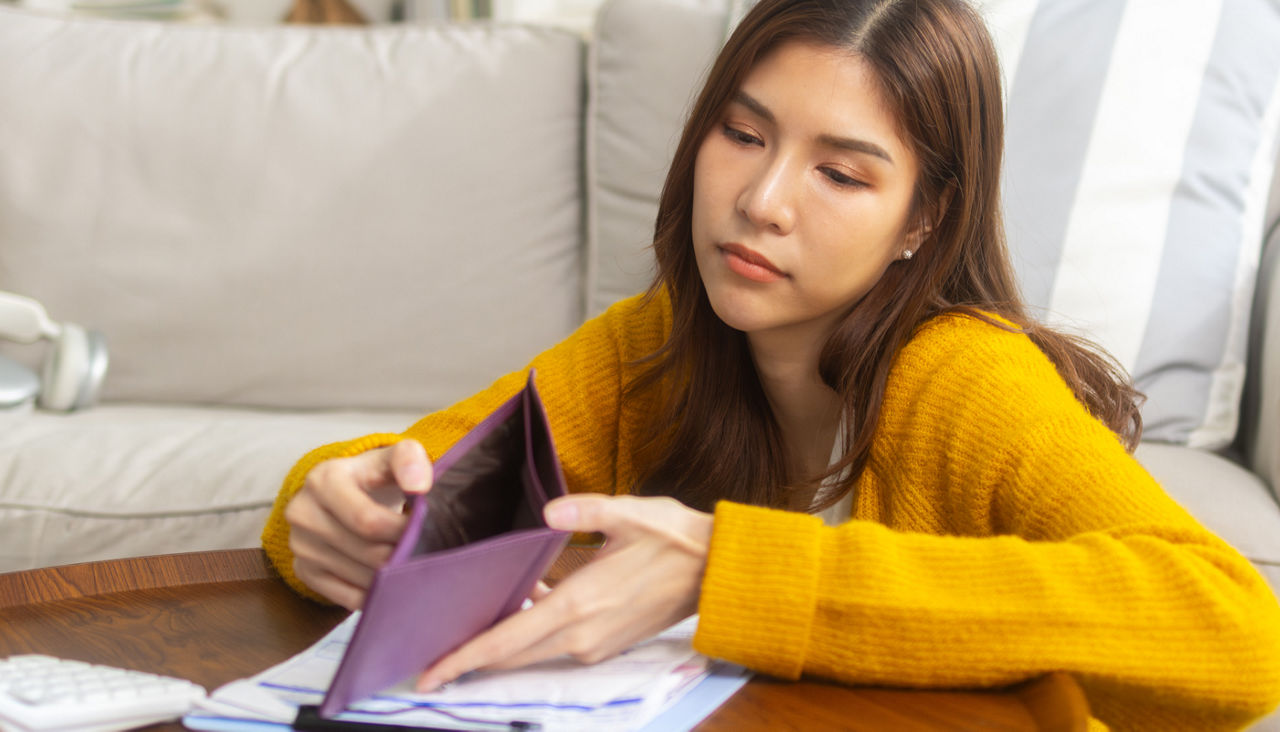 Young woman sits on floor by sofa holding an empty wallet and looking sad