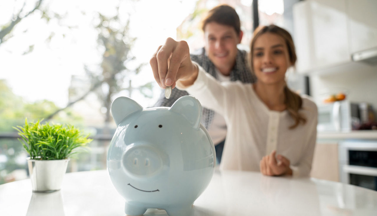 Happy couple dropping coins into a blue piggy bank on kitchen counter