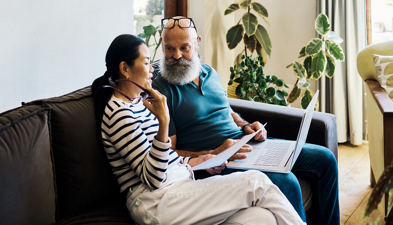 Mature couple sitting together on sofa, looking at laptop and paperwork