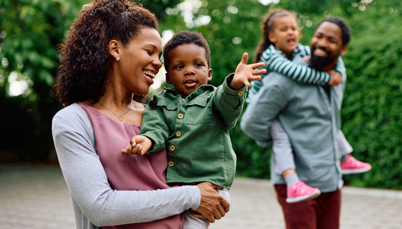 Happy African American woman holding her small son while walking outdoors. Father and daughter are in the background.