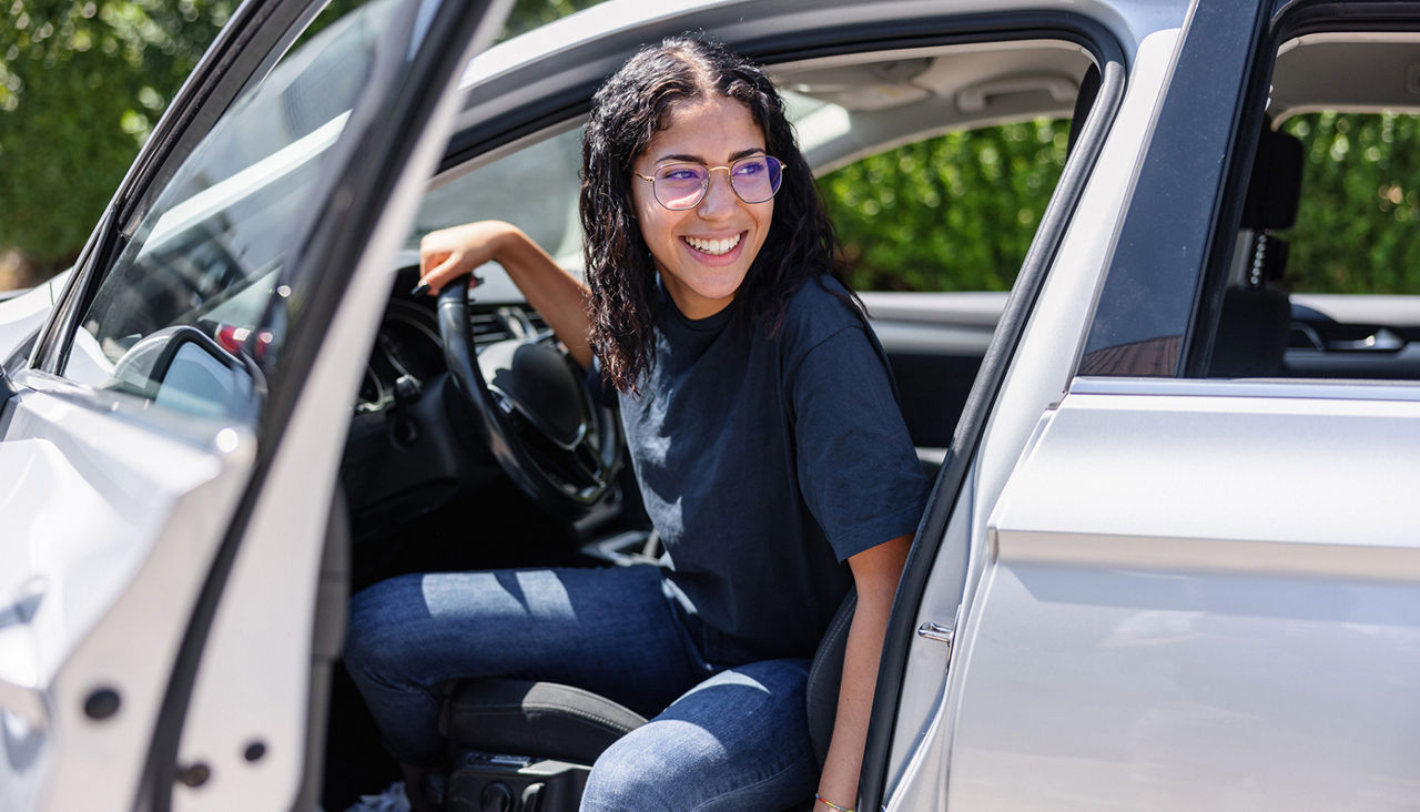 Happy female with glasses is stepping out of her parked car. The car door is open.