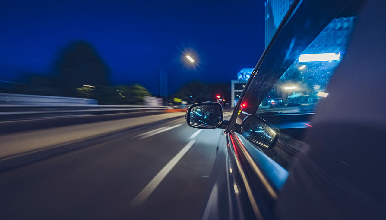 A side view of a vehicle driving along a paved roadway at night.