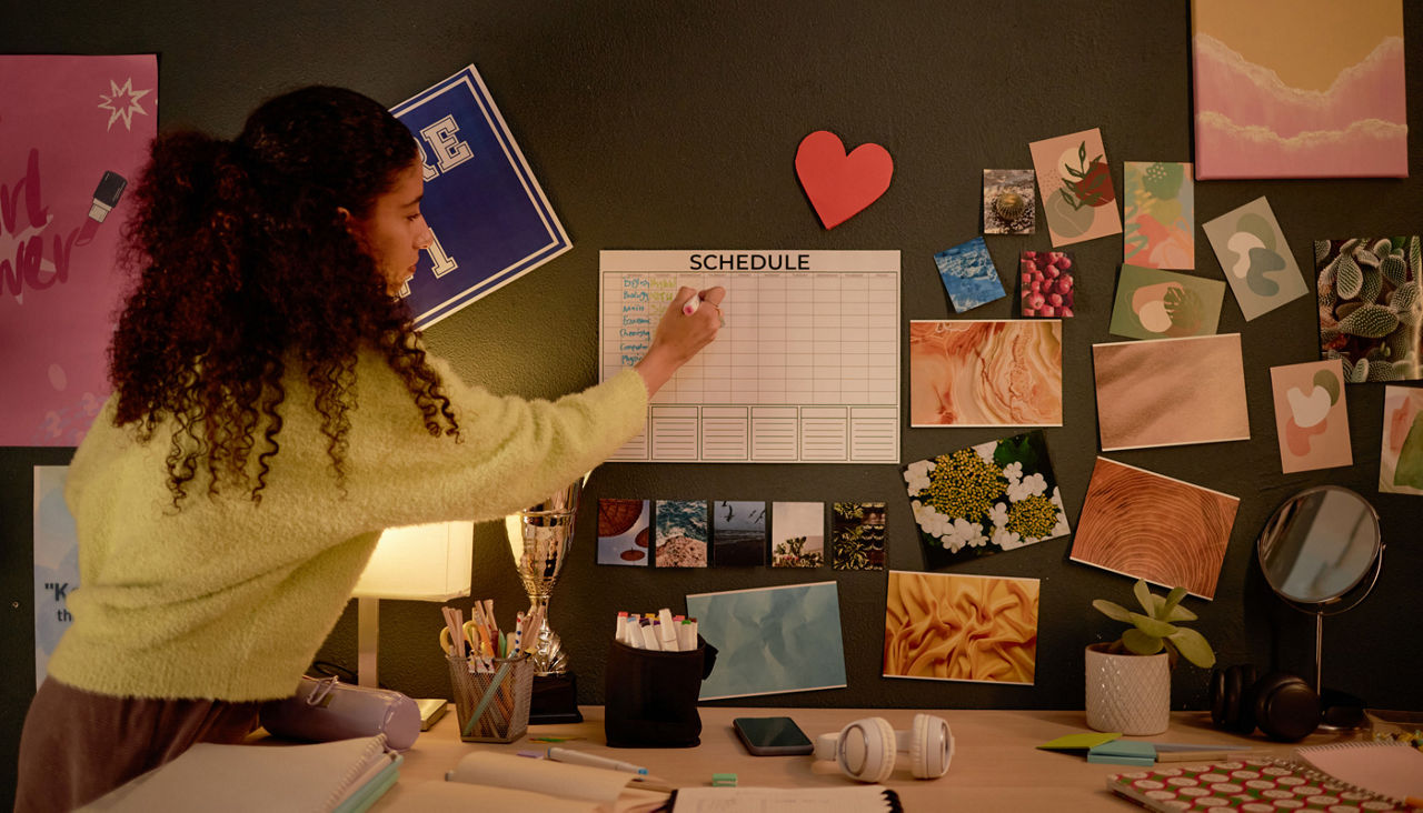 College student decorating desk with various pictures while organizing schedule on wall calendar on cluttered desk