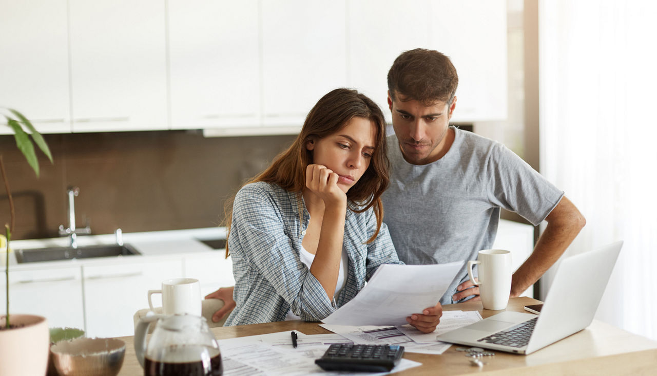 Young couple reviewing and calculating expenses in the kitchen with laptop and paperwork